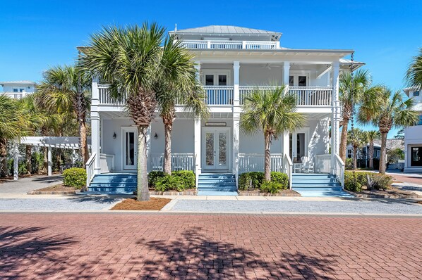 A two-story white building with a balcony, surrounded by palm trees and a brick pathway.