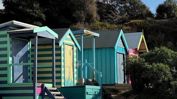A row of colorful beach huts with a sandy beach and trees in the background.