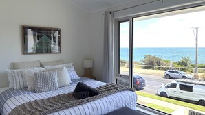 A bedroom with a large window, a bed with striped bedding, a bedside lamp, and a view of the sea and parked cars.