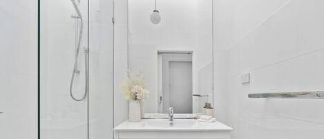 Un baño moderno con cabina de ducha de vidrio, tocador blanco con espejo y un pequeño jarrón de flores.