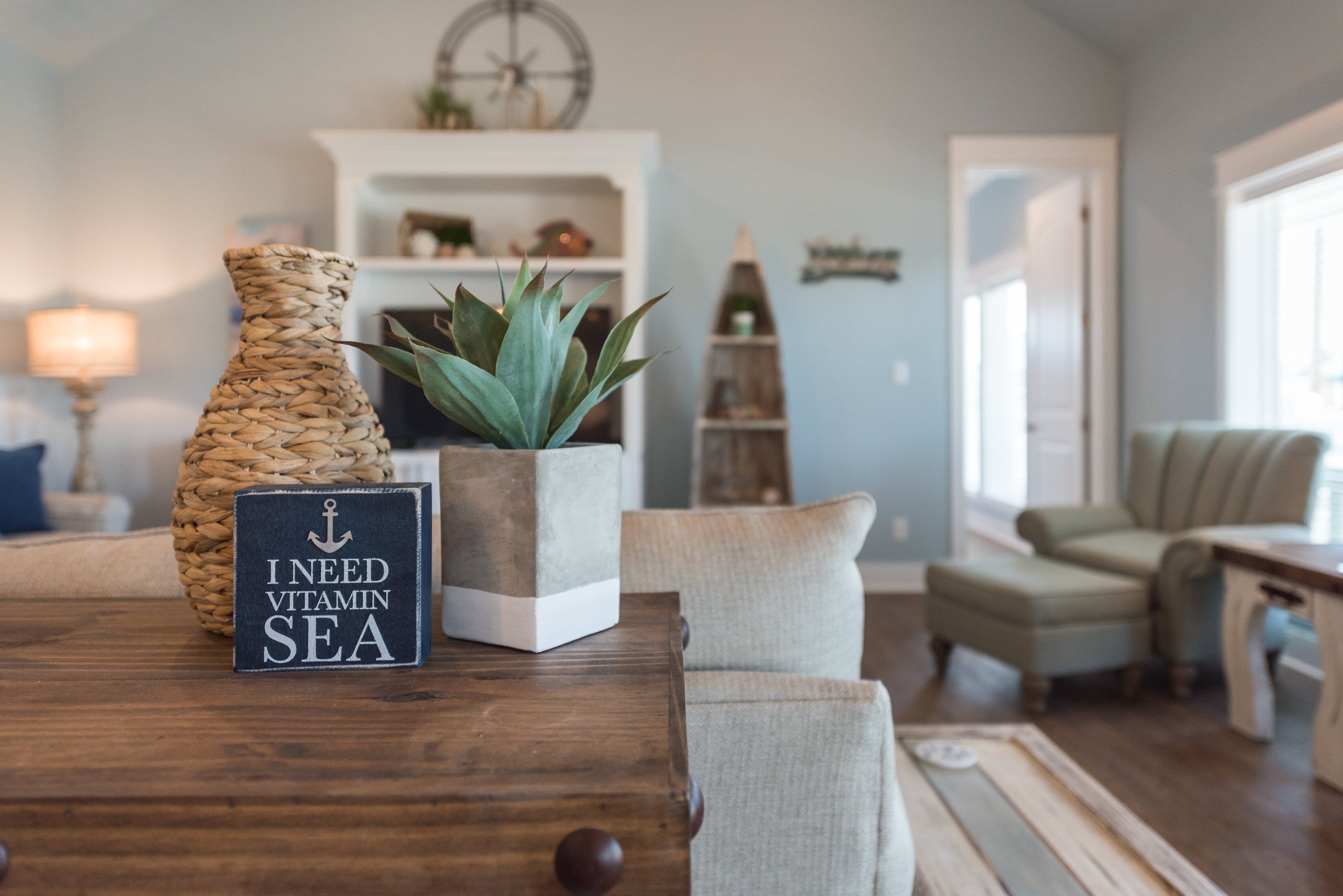 A wooden table with a sign reading "I NEED VITAMIN SEA," a woven vase, and a potted plant.