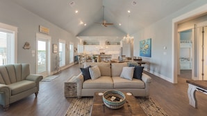 A spacious living room with a high ceiling, a ceiling fan, and a large window. - Ocean View | Modern Interior l Tiki Bar l Pet-Friendly (Crystal Beach)