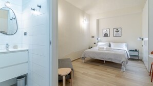 A modern hotel room with a bathroom, a bed, a small table, and a chair.