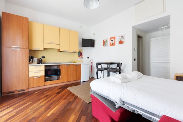 Una habitación compacta con cama, kitchenette, zona de comedor y televisor montado en la pared.
