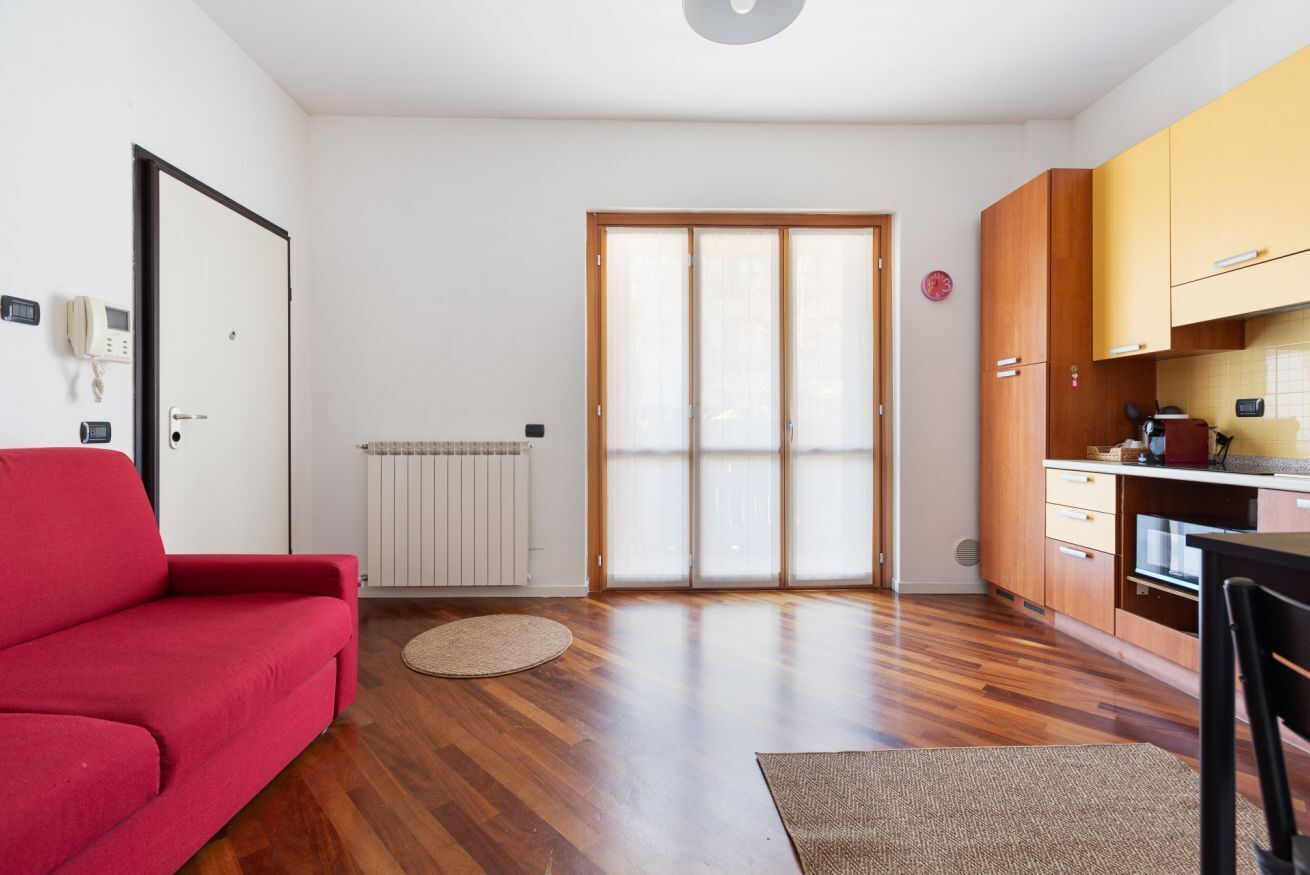 Un ambiente compatto con un divano rosso, angolo cottura con mobili in legno e una porta scorrevole in vetro che dà su uno spazio esterno.