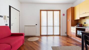 Un ambiente compatto con un divano rosso, angolo cottura con mobili in legno e una porta scorrevole in vetro che dà su uno spazio esterno.