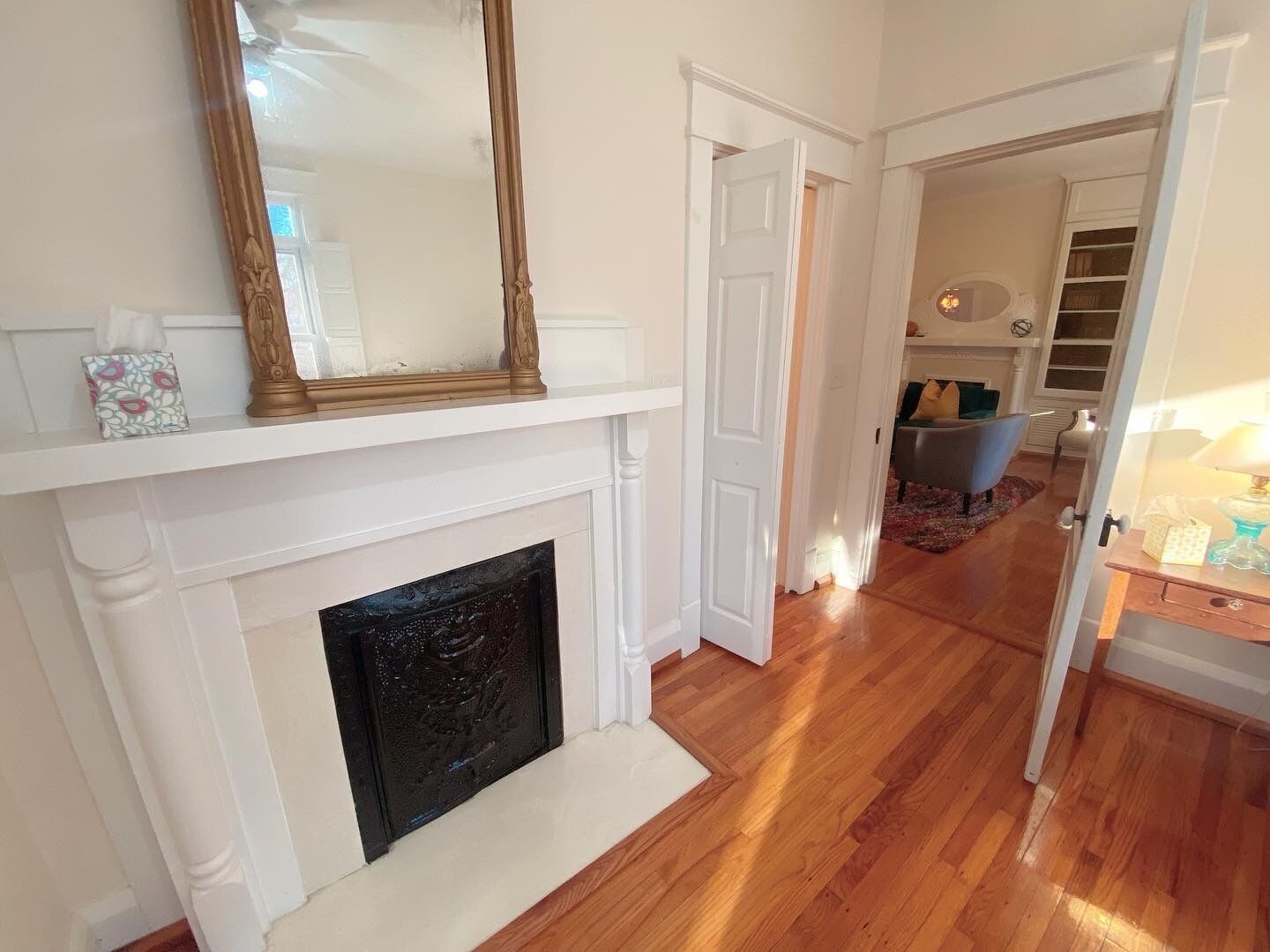 Une pièce avec une cheminée blanche, un grand miroir au-dessus et un plancher en bois.