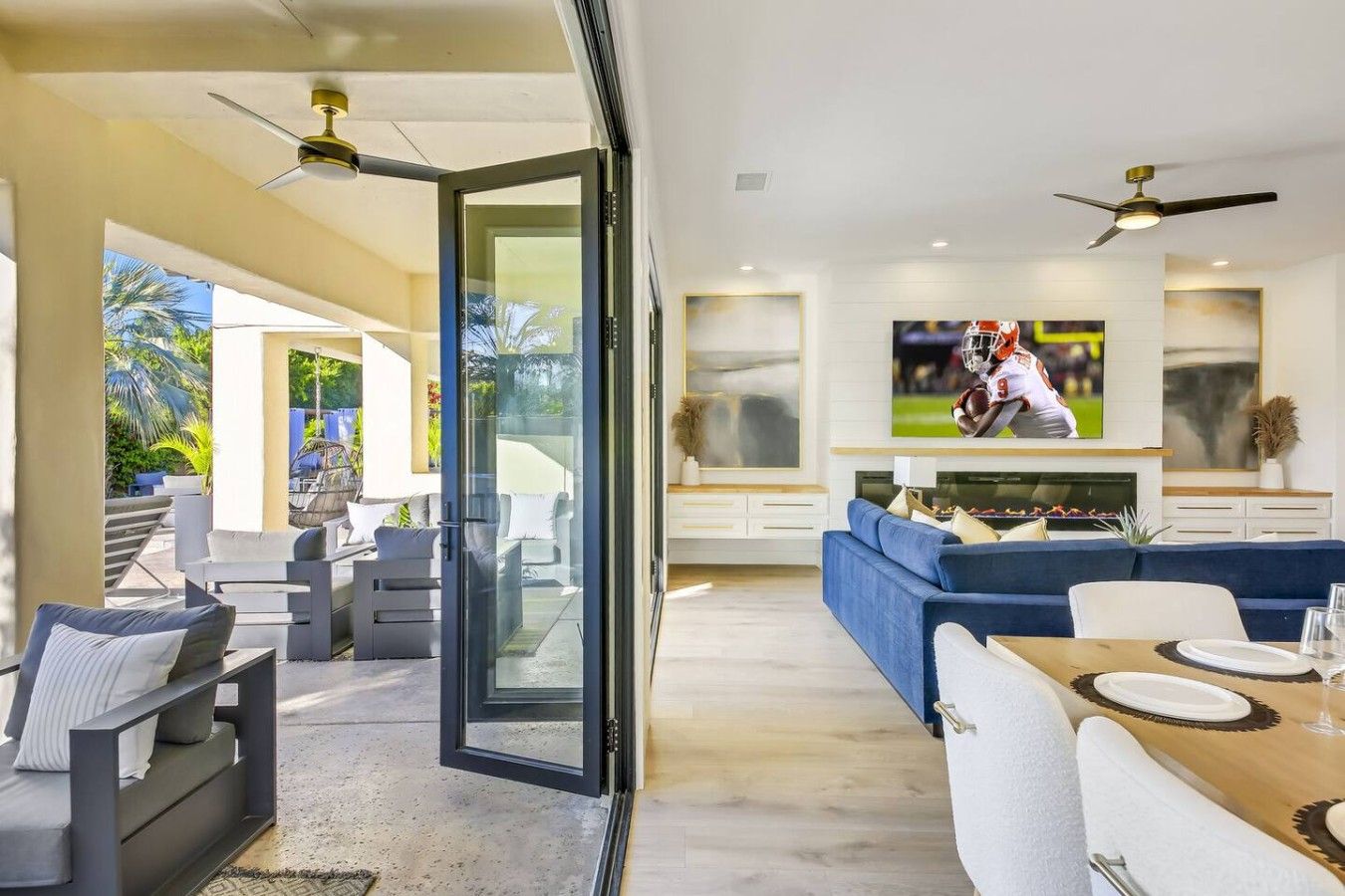 Una sala de estar moderna con un televisor grande, un comedor con una mesa lista para dos personas, y un ventanal de vidrio que da a un patio exterior.