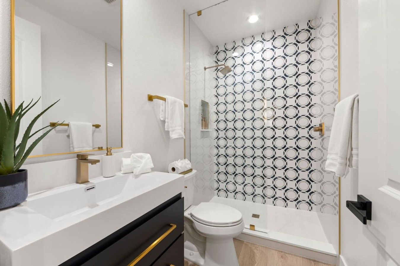 A modern bathroom with a white sink, gold fixtures, a toilet, and a shower with a geometric patterned wall.
