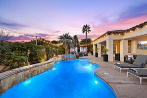 A swimming pool with lounge chairs and a covered seating area, surrounded by greenery and palm trees. - Santana: Pool, Spa, BBQ, Fire Pit, Cabanas, Games! (Indio)