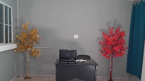 Una habitación con un escritorio, dos árboles artificiales, una ventana y una cortina azul.