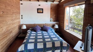 A bedroom with a wooden ceiling, a bed with a quilt, a nightstand with a lamp, a window with a view, and a television on a stand. - Cabaña con Balcón y Vista al Mar (Concón)