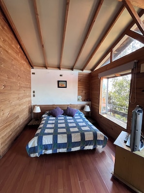 Une chambre avec un plafond en bois, un lit recouvert d’une courtepointe, une table de chevet avec une lampe, une fenêtre donnant sur un beau paysage et un téléviseur posé sur un meuble.