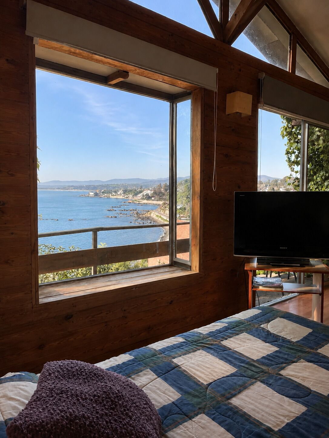 A room with a large window overlooking a coastal view, a bed with a quilt, and a television on a stand.
