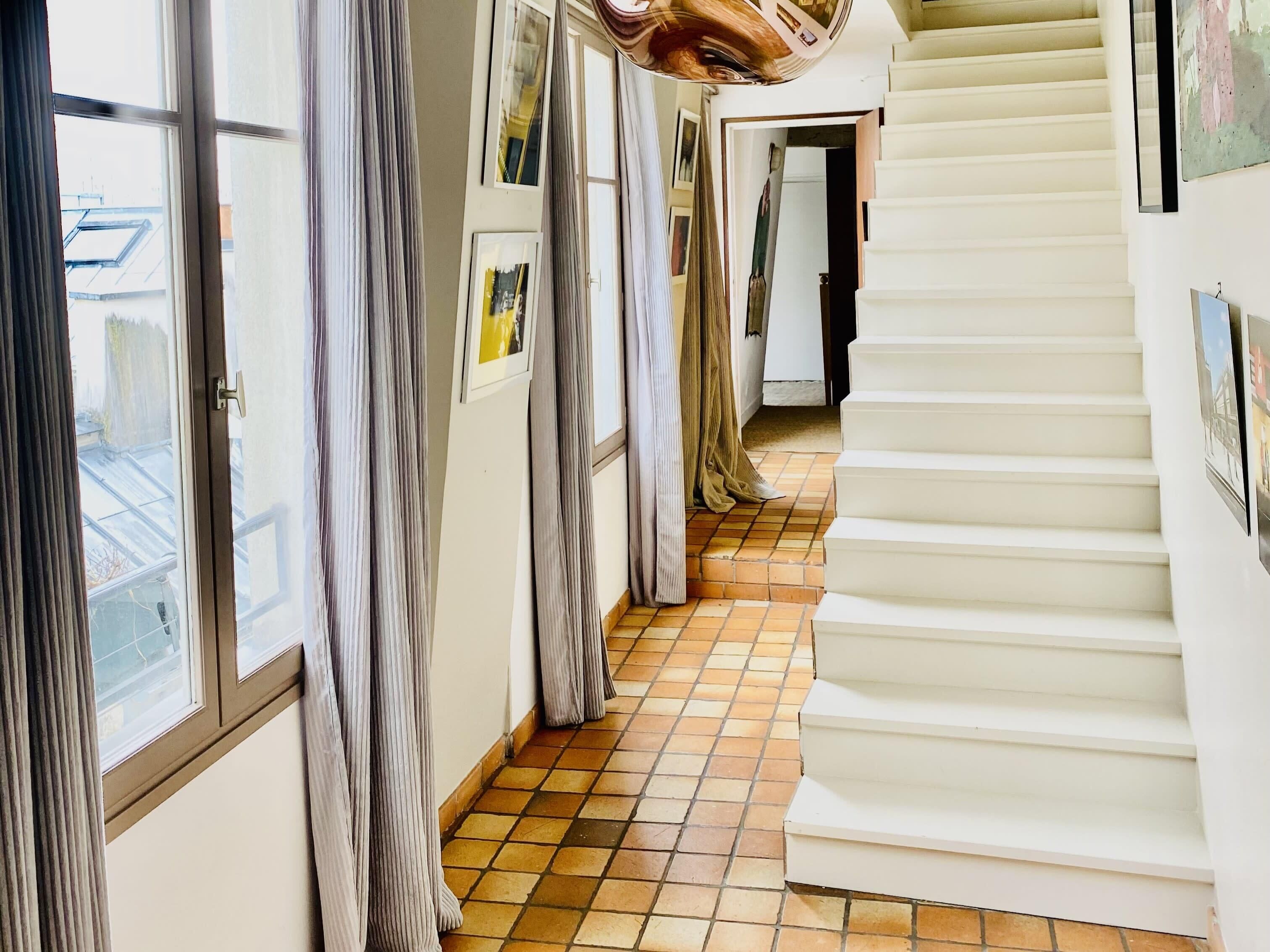 A corridor with a tiled floor, a staircase, and curtains on the windows.