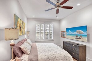 A bedroom with a bed, a ceiling fan, a window with blinds, a dresser, and artwork on the walls.