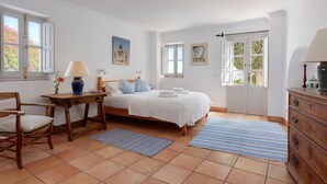 A bedroom with a bed, a wooden dresser, a chair, a table with a lamp, and a window with a view of trees. - 4 bedrooms, sea views, and pure relaxation in Frigiliana. (Frigiliana)