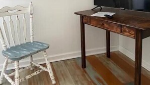Ein Zimmer mit Schreibtisch aus Holz, einem weißen Stuhl, einem großen Landschaftsbild und einem Flachbildfernseher.
