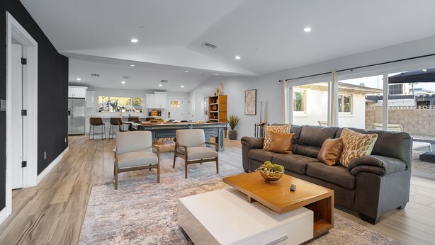 Un salon moderne avec un canapé, une table basse et des fauteuils. On aperçoit à l’arrière-plan un coin cuisine avec des meubles blancs et une table à manger.