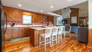 A modern kitchen with wooden cabinetry, a central island with bar stools, a dining area, and a living room with a sofa and TV. - Lost Options-Premier Home w/ Tidal Creek Dock; Creek/Marsh Views; Easy Beach Access (Edisto Beach)