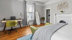 A bedroom with a bed, a desk with a laptop, a green chair, a white fireplace, and a window with curtains. - 1 Bed Luxury Downtown Columbia (Elmwood Park) (Columbia)