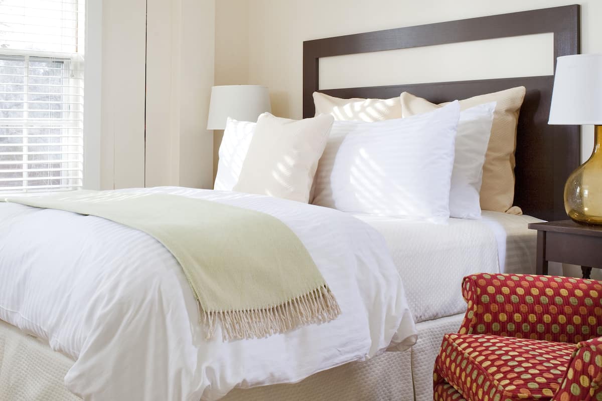 A neatly made bed with white linens and a beige throw blanket, a wooden headboard, two bedside lamps, and a patterned armchair.