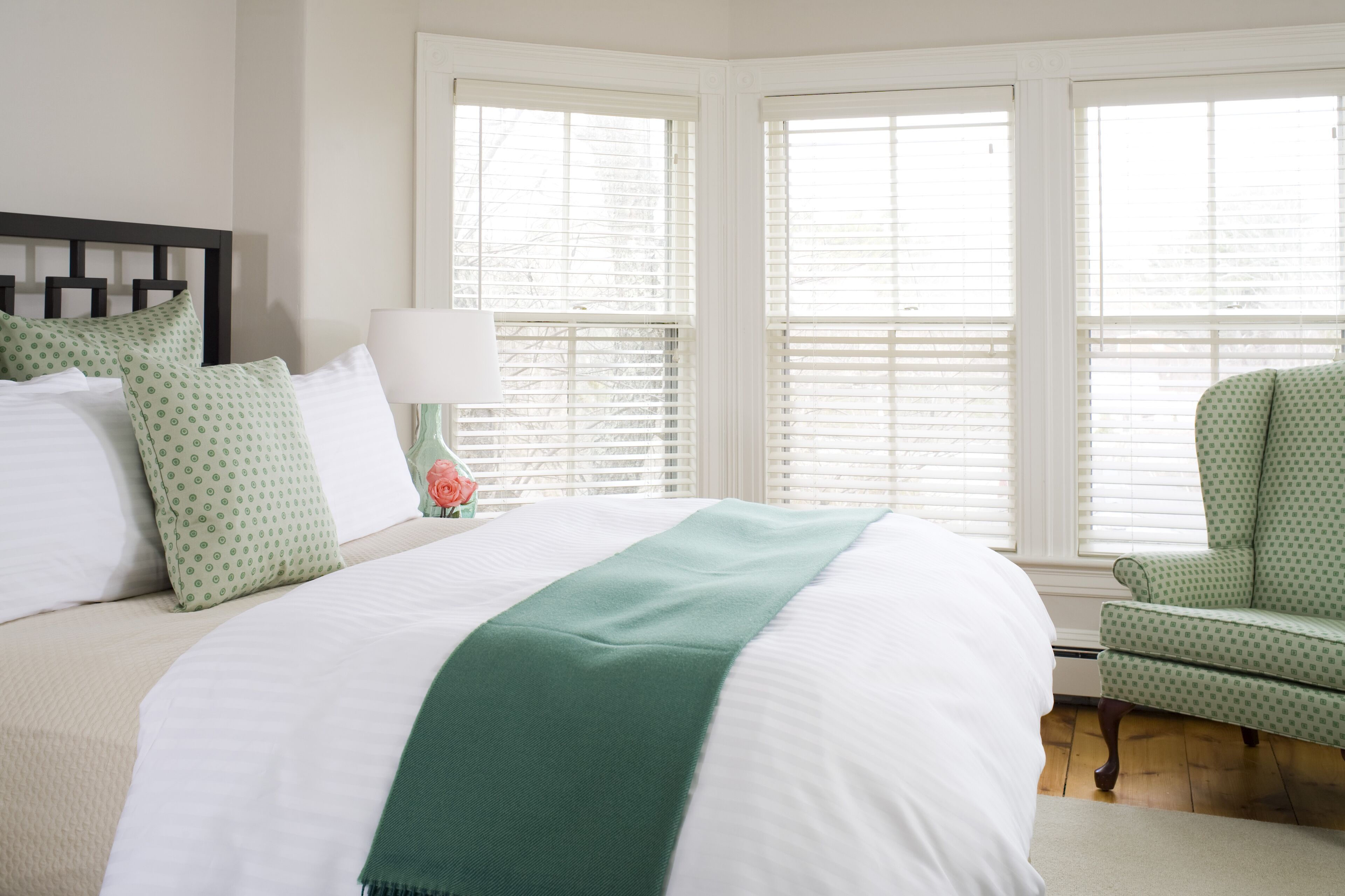 A neatly made bed with white and green bedding, a green chair, and a window with blinds.