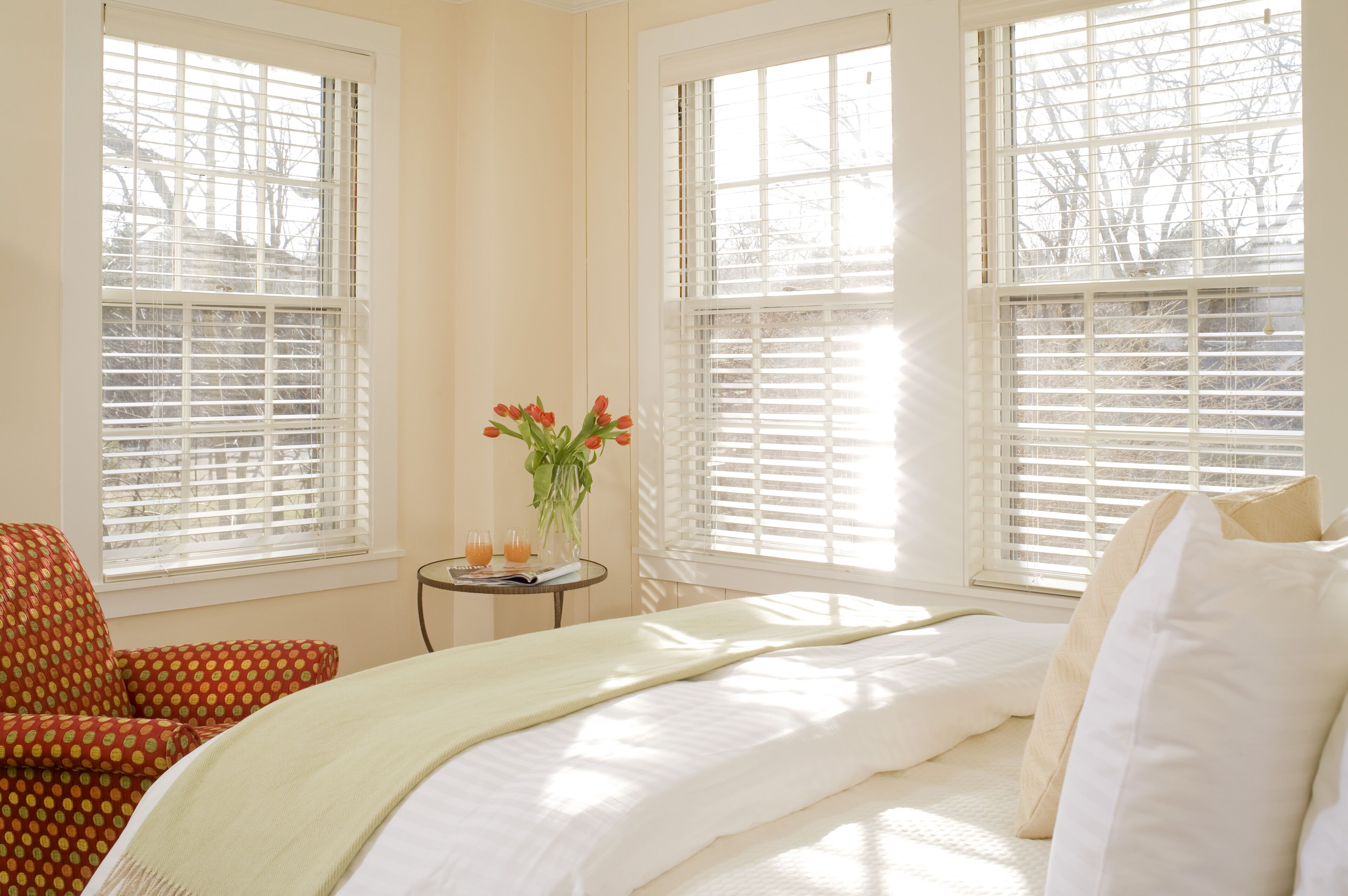A bedroom with a bed, a chair, a small table with a vase of flowers, and two windows with blinds.