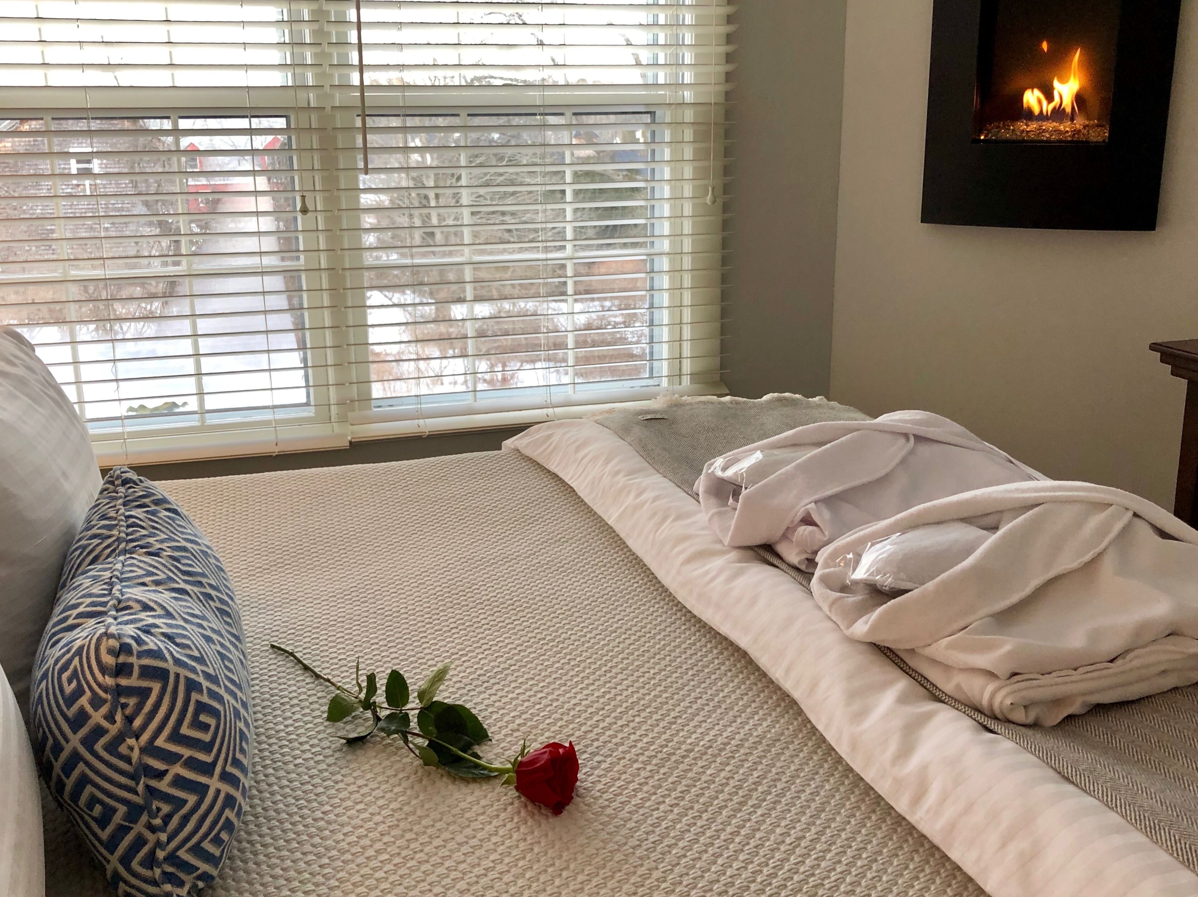 Una cama bien hecha con una manta blanca, una rosa roja solitaria y una almohada con estampado azul y blanco.