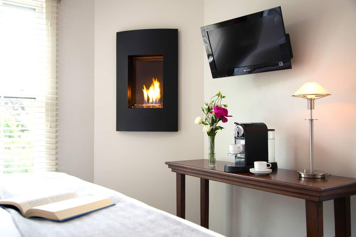 A hotel room with a bed, a bedside table with a coffee maker, a vase with flowers, a flat-screen TV, and a fireplace.