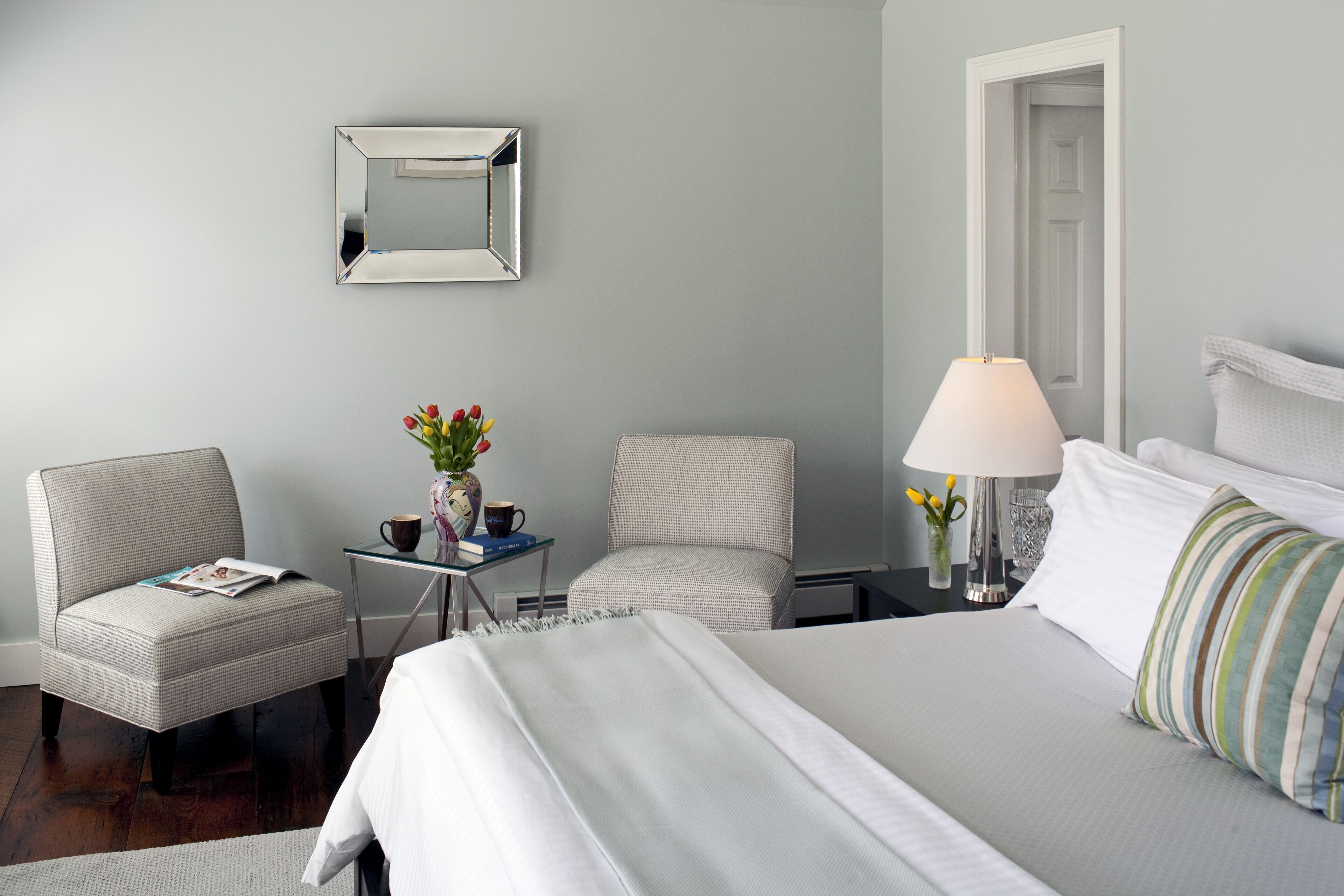 A hotel room with a bed, two chairs, a small table with a vase and books, a lamp, and a mirror on the wall.