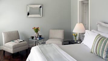 A hotel room with a bed, two chairs, a small table with a vase and books, a lamp, and a mirror on the wall.