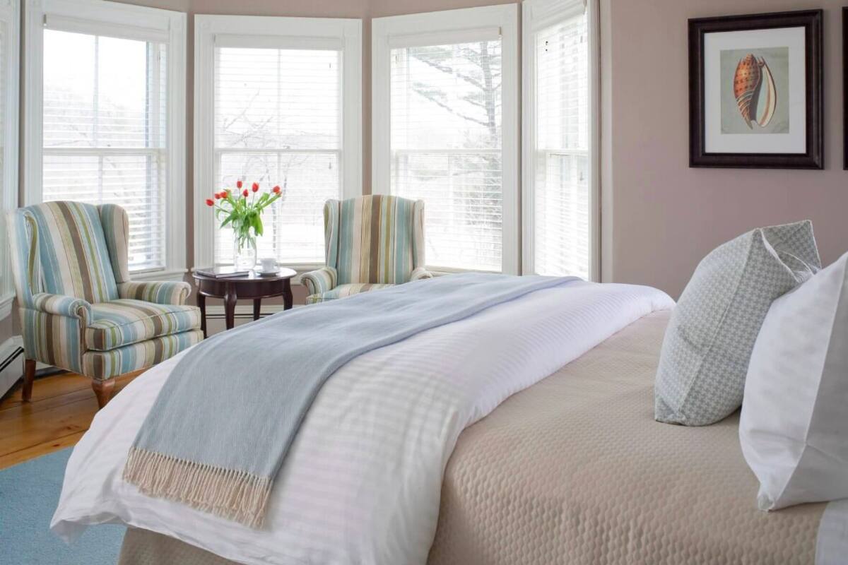 A bedroom with a large bed, two striped armchairs, a small table with a vase of flowers, and a framed picture on the wall.