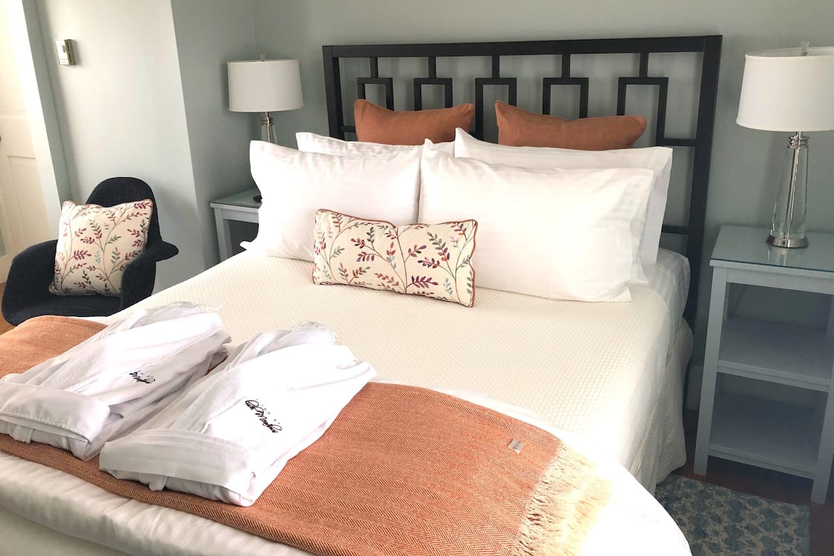 A neatly made bed with white linens, a brown throw blanket, and multiple pillows including floral and solid colors. There are two bedside tables with lamps, a chair with a floral pillow, and a small table with a glass vase.
