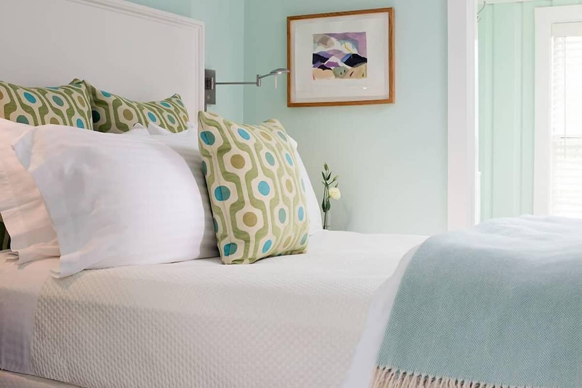 A neatly made bed with white and patterned pillows, a light blue herringbone throw blanket, and a framed artwork on the wall.