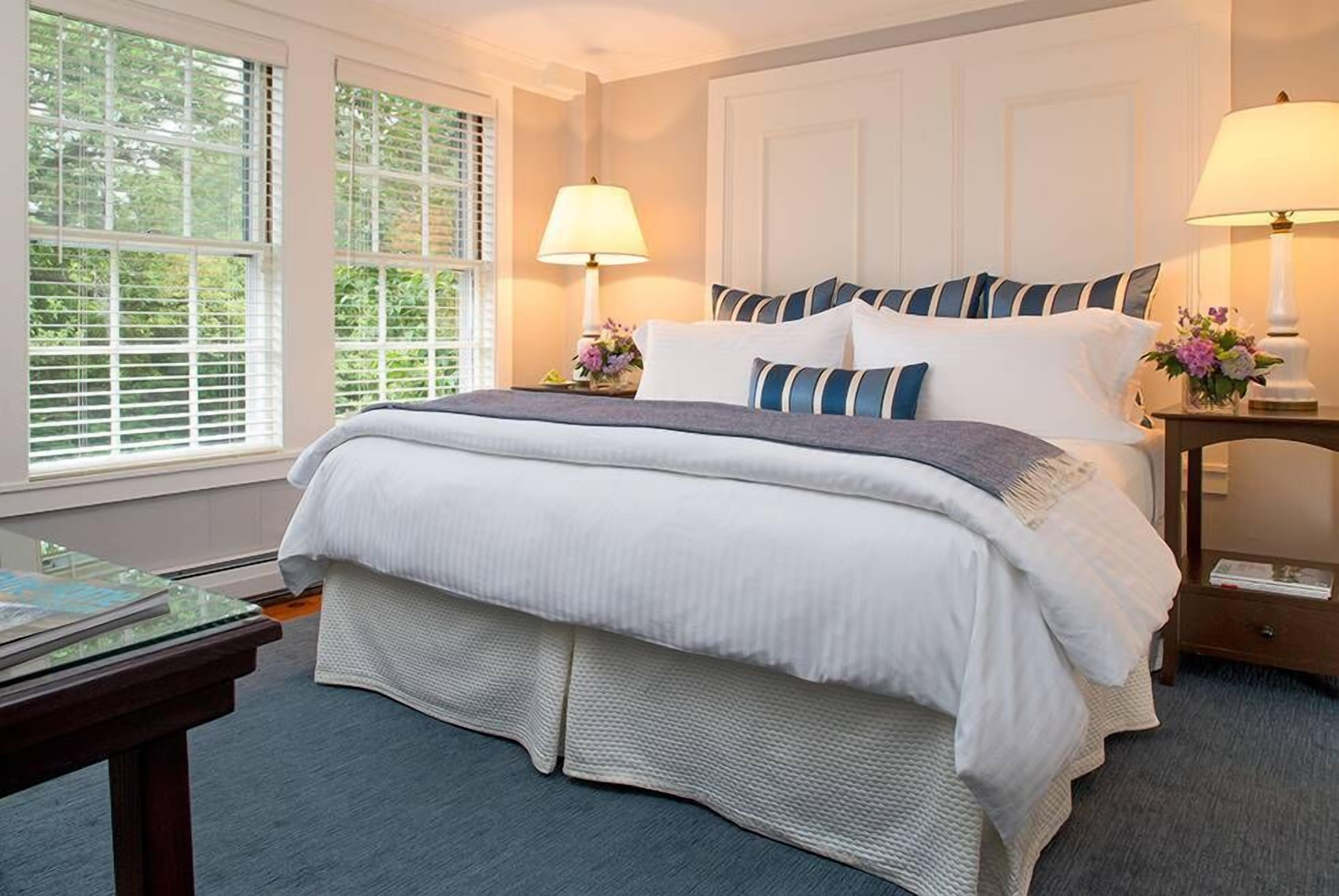 A neatly made bed with white and blue striped pillows, two bedside lamps, and a window with blinds.