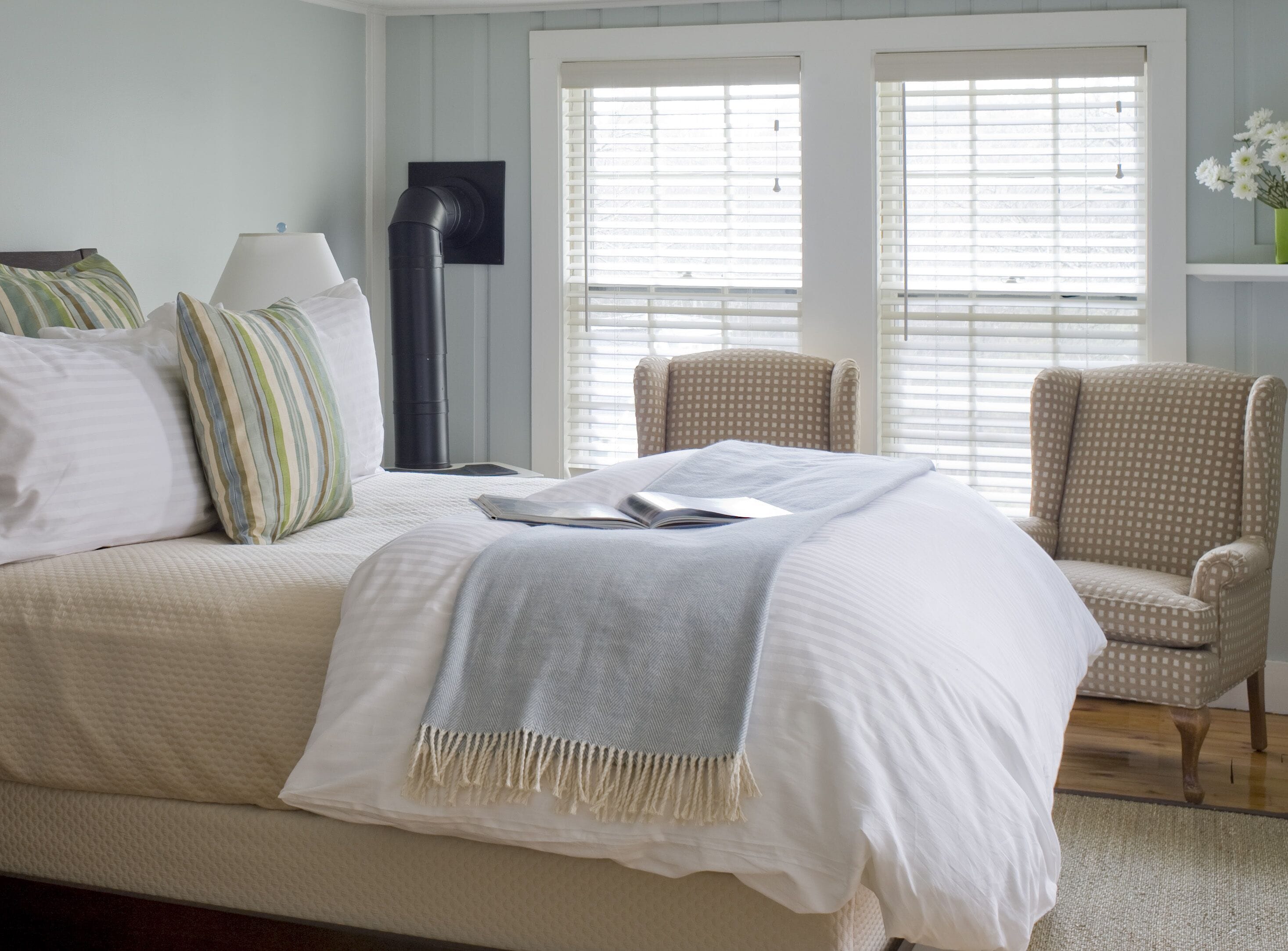 A neatly arranged bedroom with a large bed, two chairs, a standing lamp, and a window with blinds.