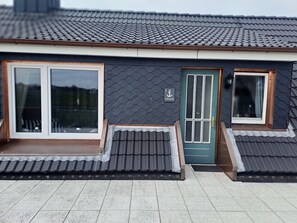 Un bâtiment avec un toit recouvert de tuiles sombres, une porte verte et une fenêtre dotée d’un cadre blanc.