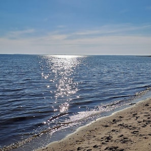 A beach with calm water reflecting sunlight, a clear sky, and a distant shoreline.