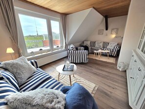 A loft-style room with a sofa, armchair, and small table. A window offers a view of a garden. - Haus Dorfschmiede, Whg. Ankerplatz - Haus Dorfschmiede (Alkersum)