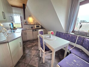 A compact kitchen with a microwave, kettle, and sink. There is a dining table with a vase of flowers and purple upholstered seating.