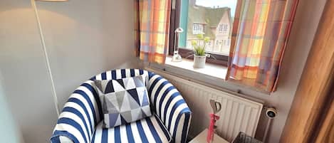Una habitación de esquina con un sillón a rayas, una mesita con una cesta, un radiador y una ventana con cortinas.