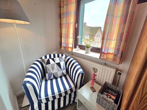 Une pièce d’angle avec un fauteuil à rayures, une petite table sur laquelle se trouve un panier, un radiateur et une fenêtre avec des rideaux.