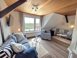 A loft-style room with a sloped ceiling, a large window with a view, a striped sofa, a striped armchair, and a small round table.
