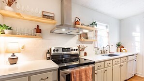 Cocina con armarios blancos, estufa y pileta. Se observan estantes con cristalería y una planta.