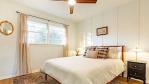 Un dormitorio con cama, ventilador de techo, ventana con cortinas, mesita de noche con lámpara y un cuadro en la pared.