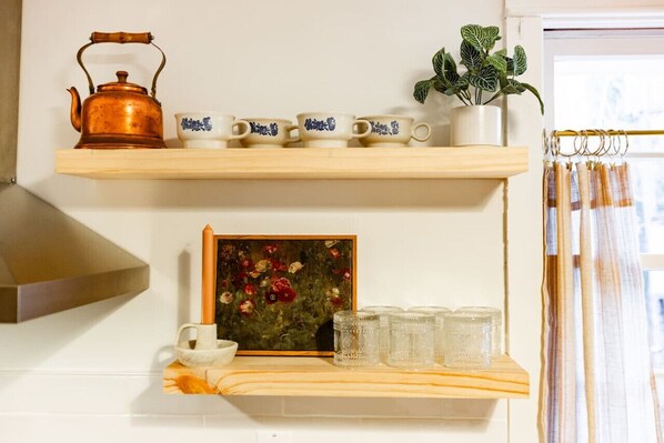 A kitchen shelf with a copper kettle, white cups with blue designs, a framed painting, and clear glass jars.