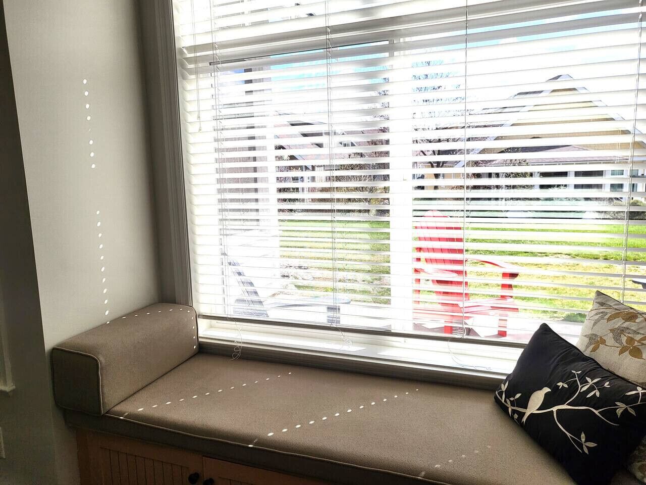 A window seat with a cushion, a view of a red chair outside, and a window with blinds.