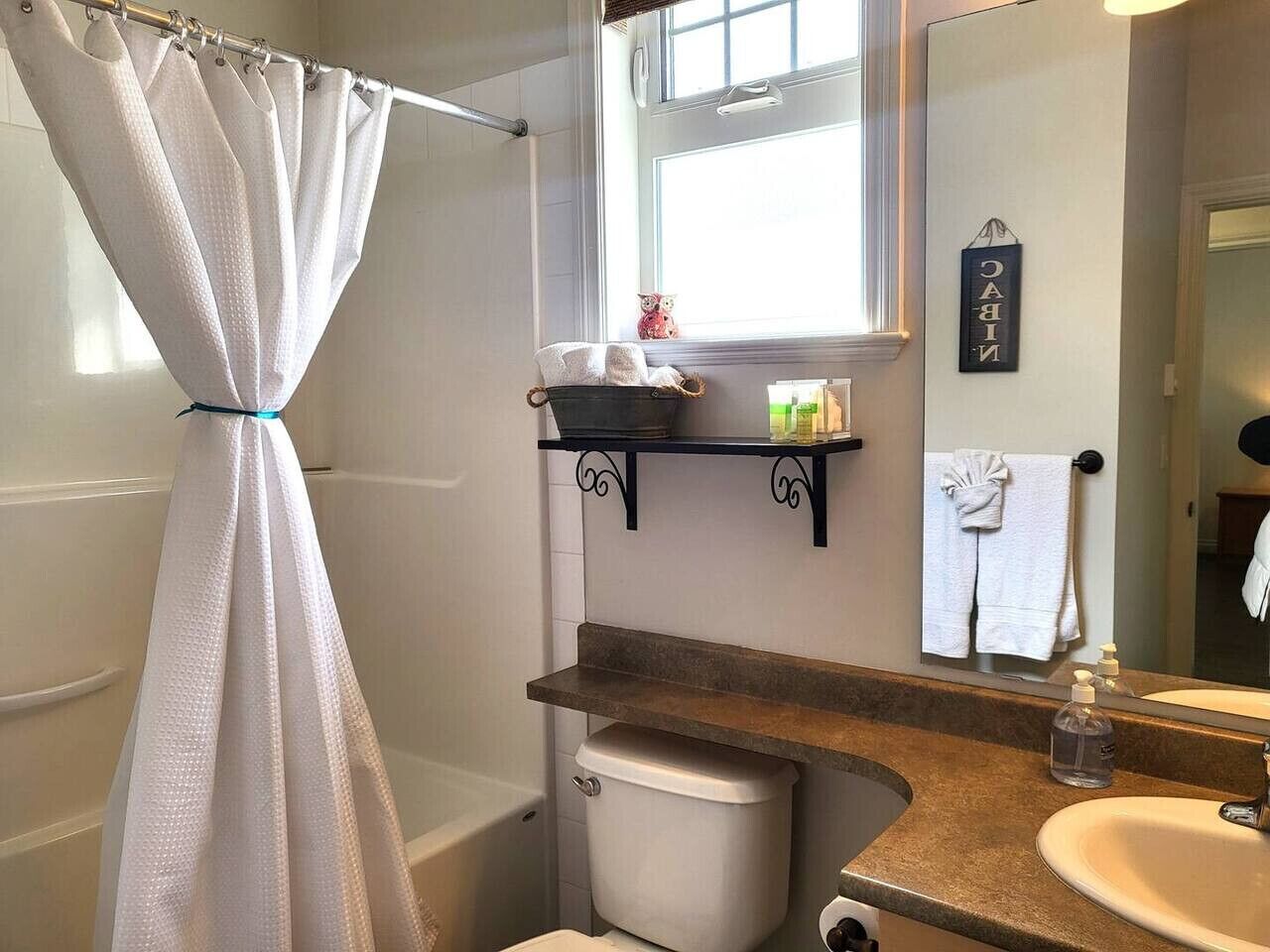 Et badeværelse med bruseforhæng, toilet, vask, spejl og et vindue.