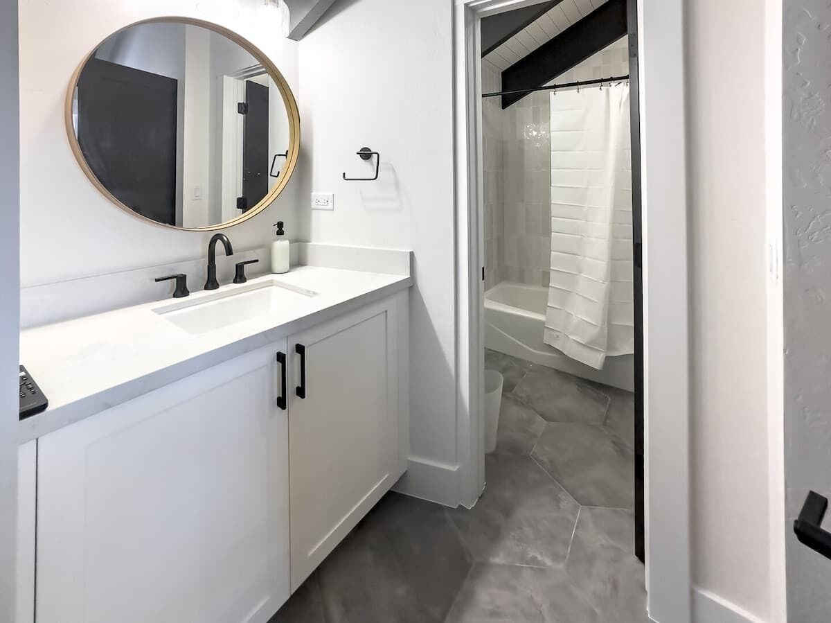 Une salle de bain avec un meuble-lavabo blanc, un miroir rond, un robinet noir et un espace douche avec un rideau blanc.
